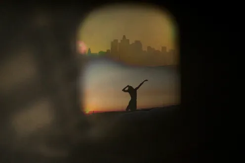Silhouette of single dancer in front of a city-scape, seen through a window surrounded by darkness