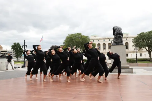 Group of dancers dressed in black with arched posture each with a hand on their forehead. 