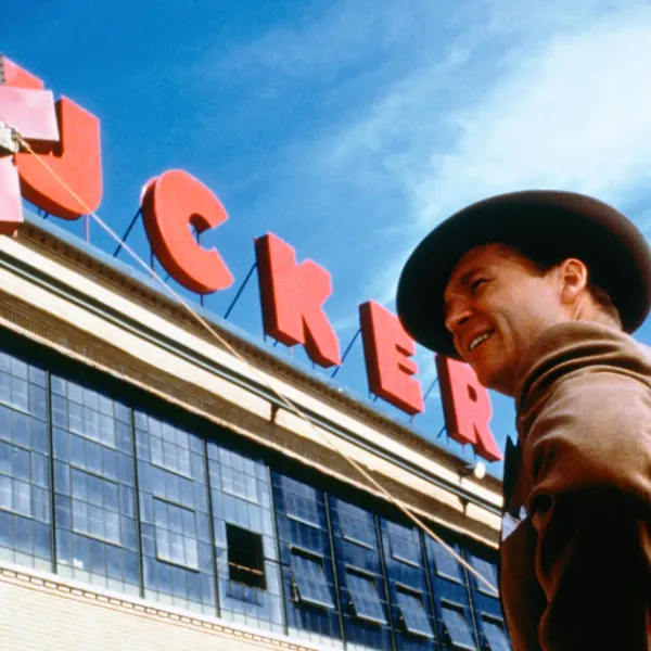 A man with a wide brimmed hat stands below a large red sign reading "Tucker" as it is assembled.