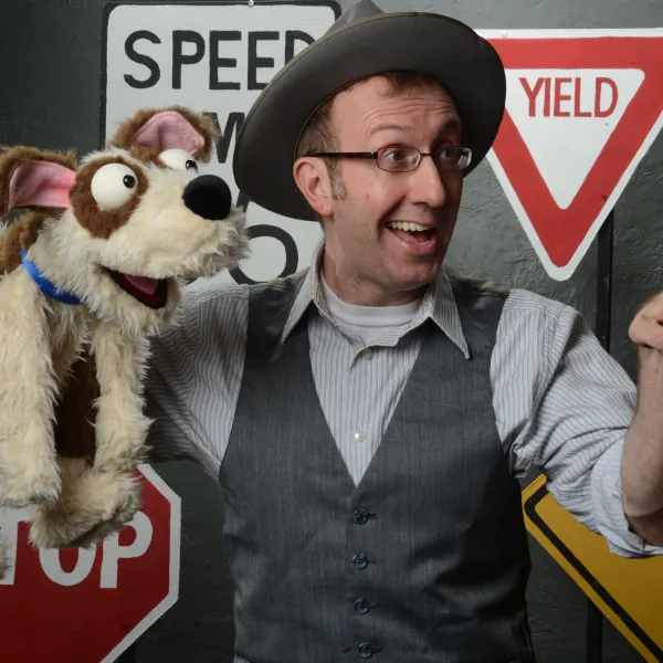 A man in a wide brimmed hat and vest holds a dog puppet in one hand and points out of view with the other. Various road signs are assembled behind him.