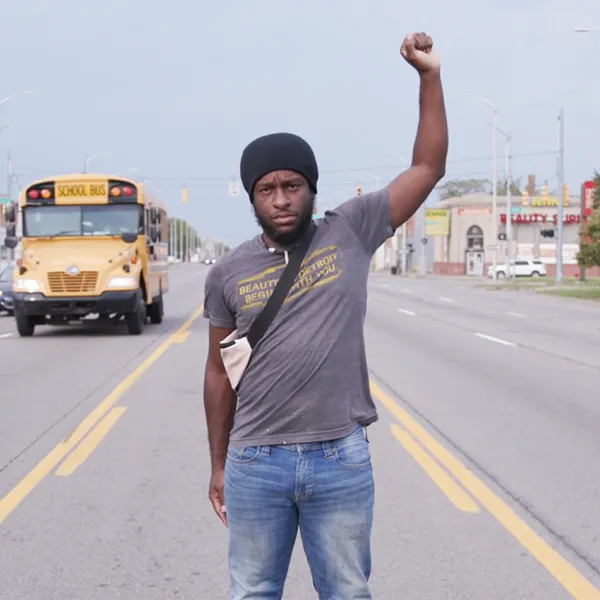 A Black man stands in the middle of a turn lane on a six lane highway with his left arm raised in a fist.