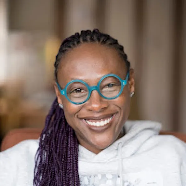 Nandi Comer, smiling with braided hair and turquoise circle-framed glasses.