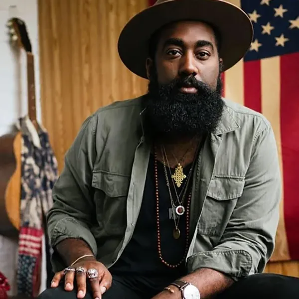 A Black man in a wide-brimmed hat sits in front of a guitar and an American flag
